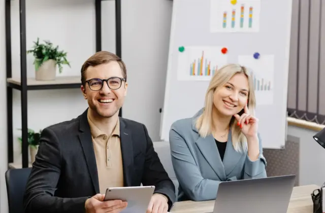 Two colleagues smiling in a meeting room with charts behind them